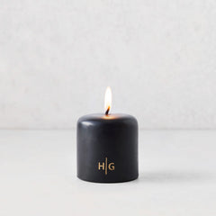 3x3 inch black hand-dipped pillar candle with a burning wick and gold embossed HG logo on a white surface.
