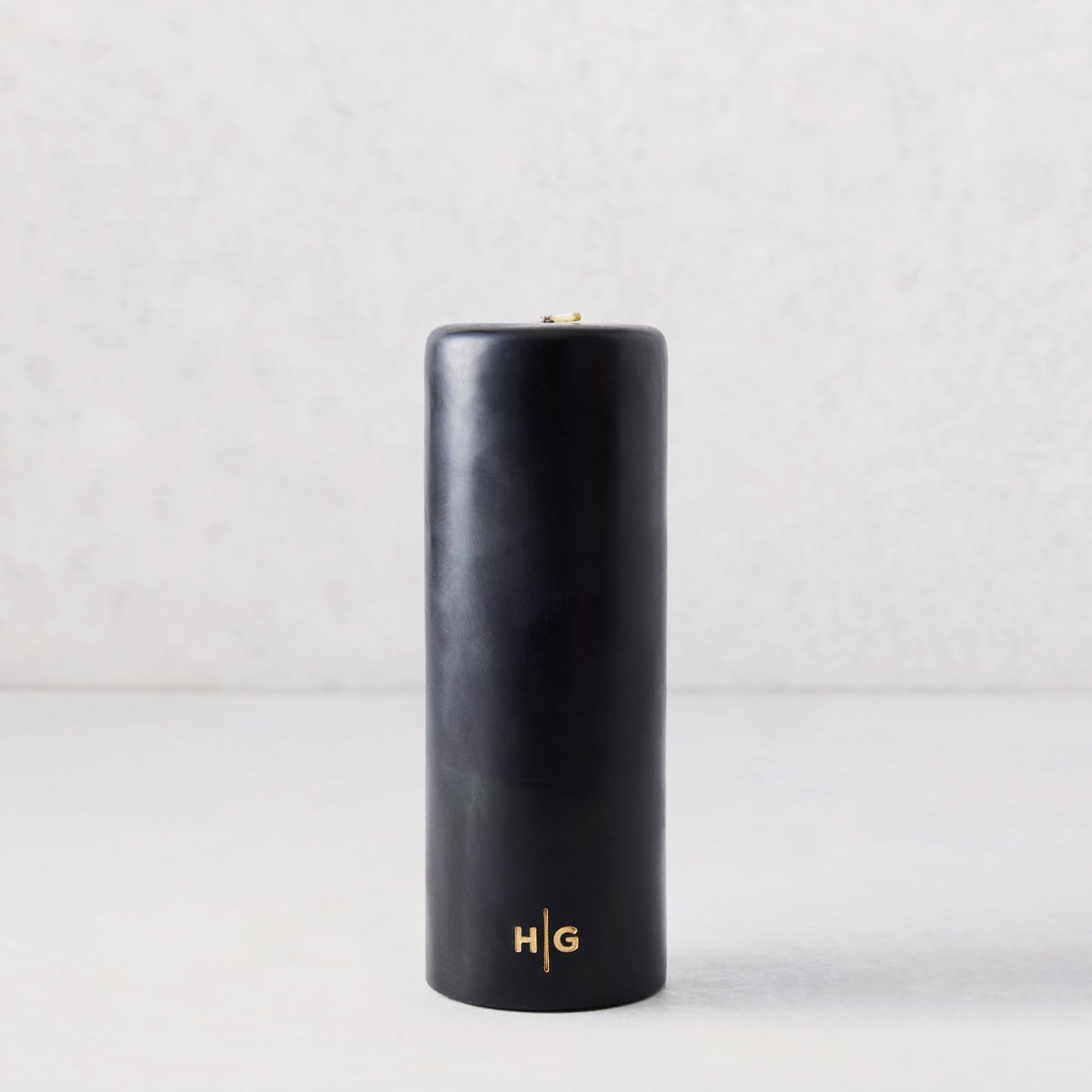3x8 inch black unscented pillar candle with a gold HG logo embossed on the base, set against a light background.
