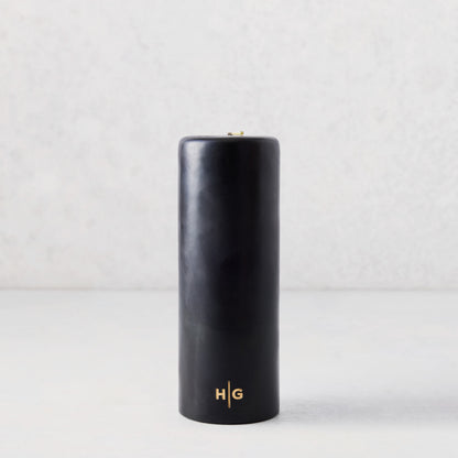 3x8 inch black unscented pillar candle with a gold HG logo embossed on the base, set against a light background.