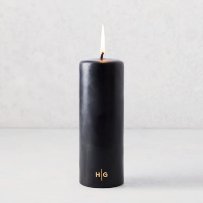 3x8 inch black unscented pillar candle with a gold HG logo and lit cotton wick.