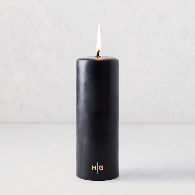 3x8 inch black unscented pillar candle with a gold HG logo and lit cotton wick.