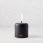 4"x4" black unscented pillar candle with a burning cotton wick and gold embossed HG logo on a light background.