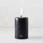 4x6 inch black unscented pillar candle with a burning wick and gold HG logo on a light background.