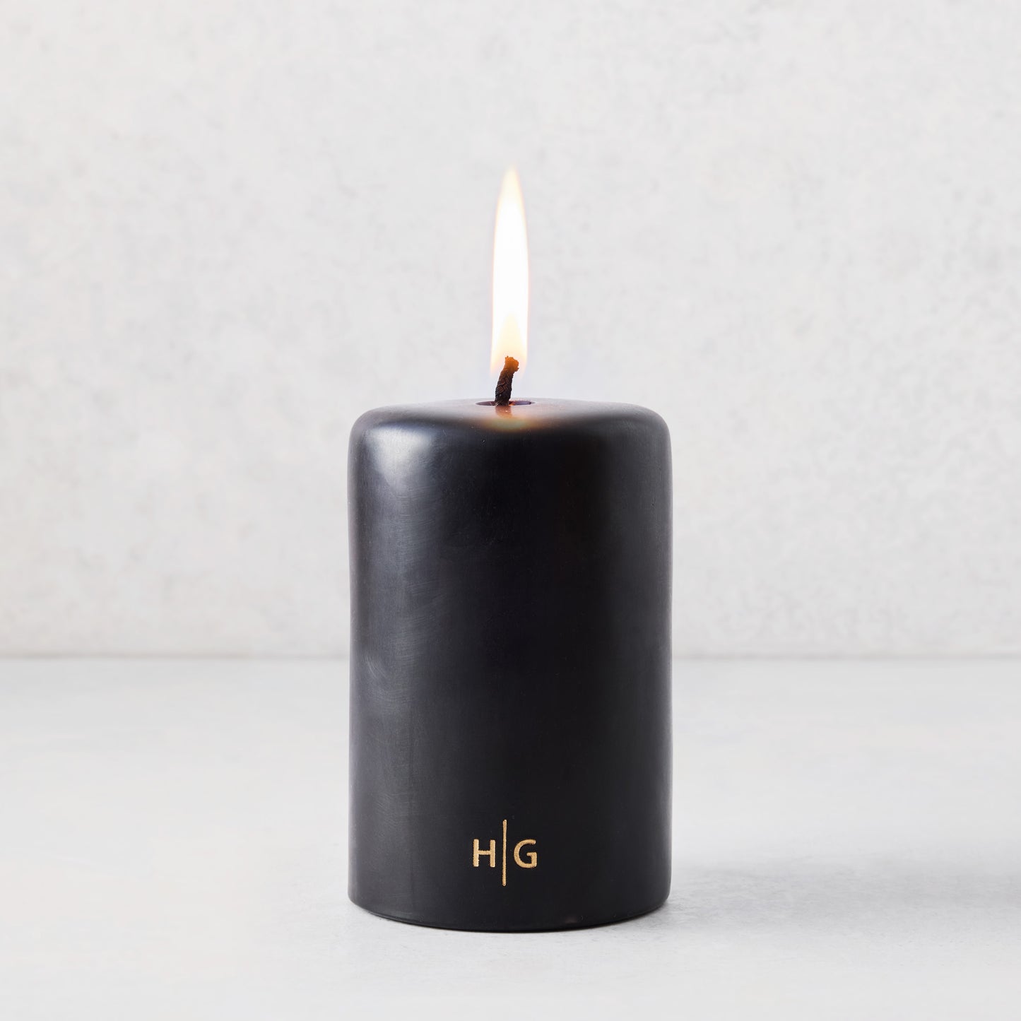 4x6 inch black unscented pillar candle with a burning wick and gold HG logo on a light background.