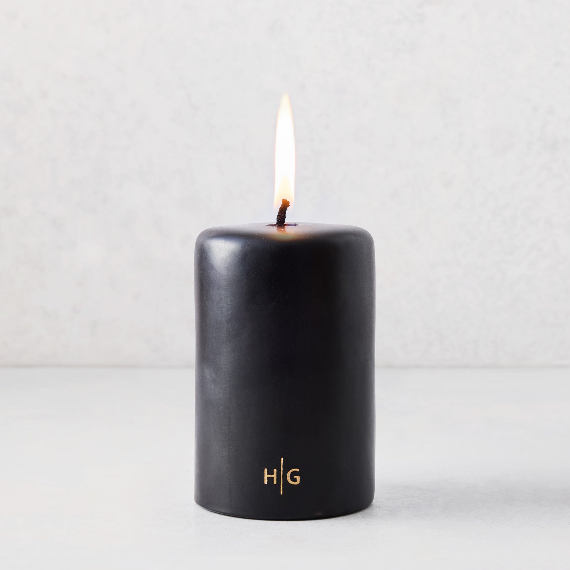 4x6 inch black unscented pillar candle with a burning wick and gold HG logo on a light background.