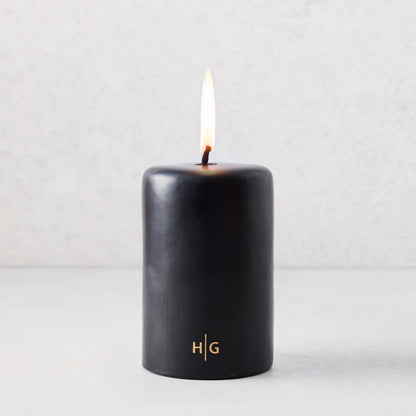 4x6 inch black unscented pillar candle with a burning wick and gold HG logo on a light background.