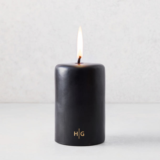 4x6 inch black unscented pillar candle with a burning wick and gold HG logo on a light background.