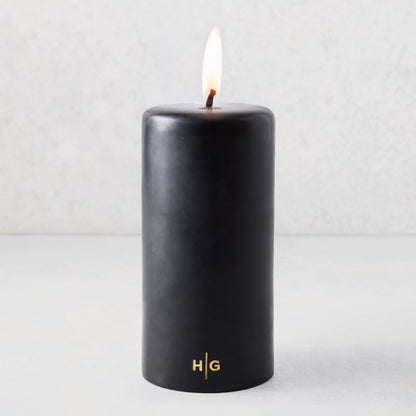 4x8 inch black unscented pillar candle with a burning wick and gold HG logo embossed on the base.