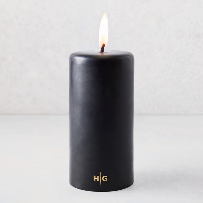 4x8 inch black unscented pillar candle with a burning wick and gold HG logo embossed on the base.