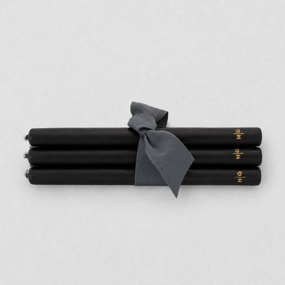 Four black taper candles tied together with a dark gray ribbon, featuring small gold lettering near the ends, on a light gray background.