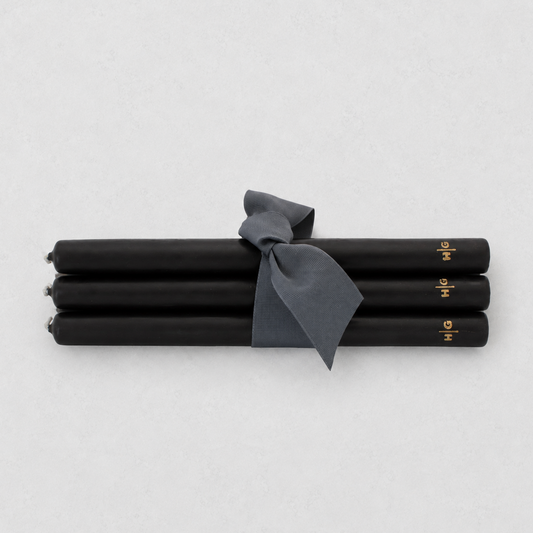 Four black taper candles tied together with a dark gray ribbon, featuring small gold lettering near the ends, on a light gray background.