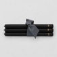 Four black taper candles tied together with a dark gray ribbon, featuring small gold lettering near the ends, on a light gray background.