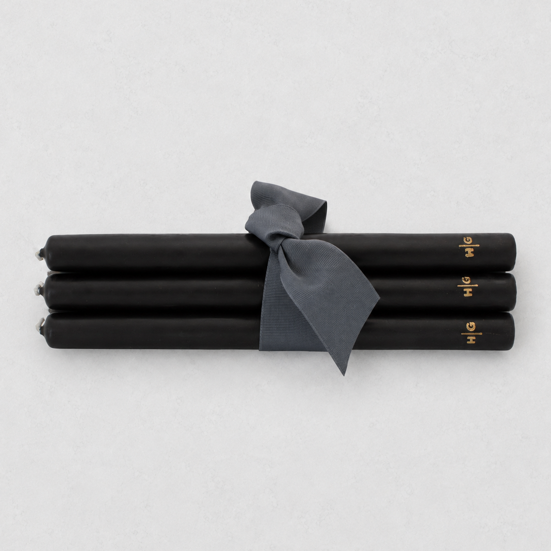 Four black taper candles tied together with a dark gray ribbon, featuring small gold lettering near the ends, on a light gray background.