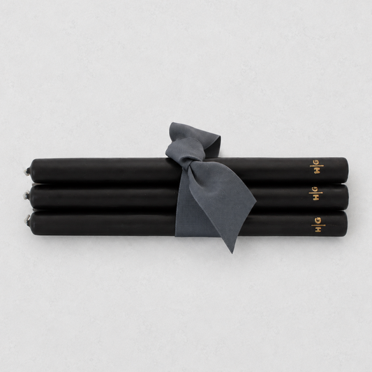 Four black taper candles tied together with a dark gray ribbon, featuring small gold lettering near the ends, on a light gray background.