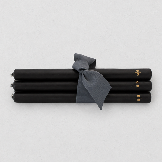 Four black taper candles tied together with a dark gray ribbon, featuring small gold lettering near the ends, on a light gray background.