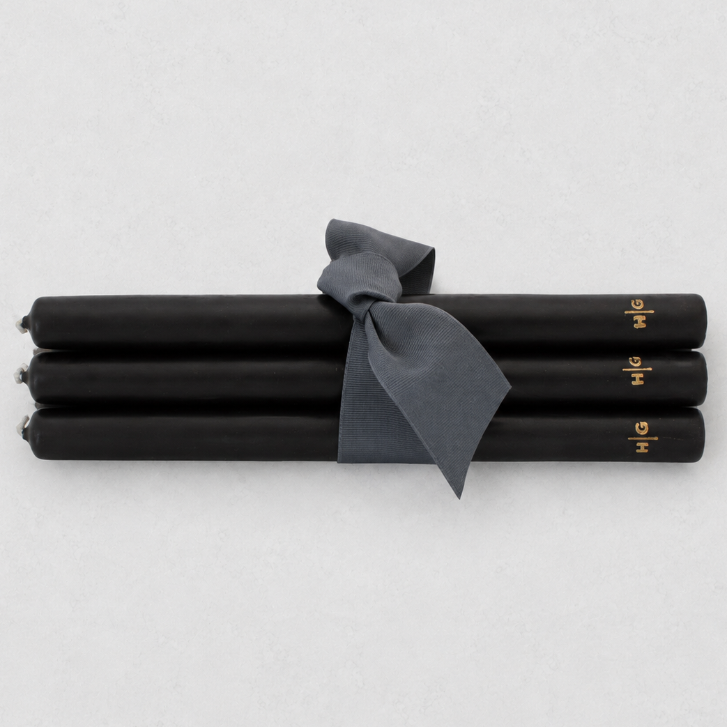 Four black taper candles tied together with a dark gray ribbon, featuring small gold lettering near the ends, on a light gray background.