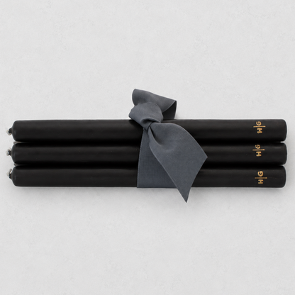 Four black taper candles tied together with a dark gray ribbon, featuring small gold lettering near the ends, on a light gray background.