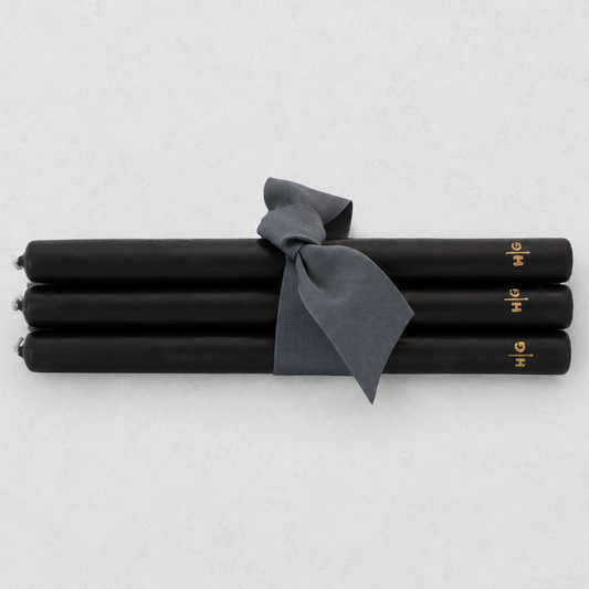 Four black taper candles tied together with a dark gray ribbon, featuring small gold lettering near the ends, on a light gray background.