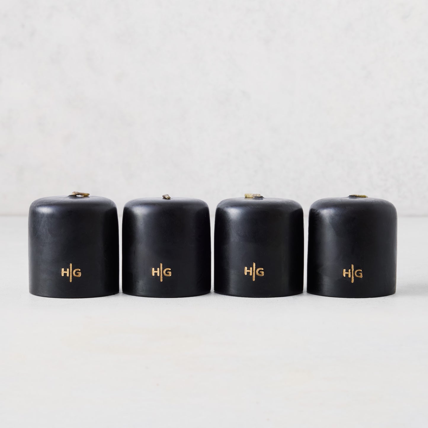 Set of four 2-inch black unscented votive candles with a gold embossed HG logo on a light background.