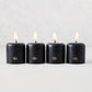 Set of four 2-inch black unscented votive candles with lit wicks and gold embossed HG logo on a light background.