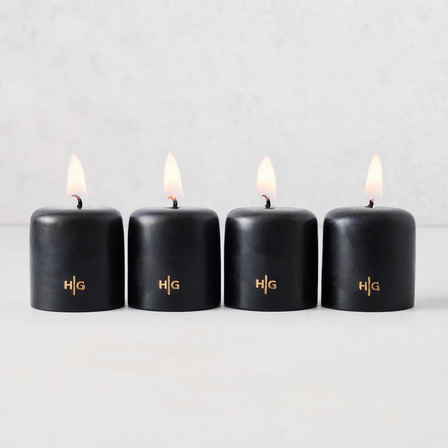 Set of four 2-inch black unscented votive candles with lit wicks and gold embossed HG logo on a light background.