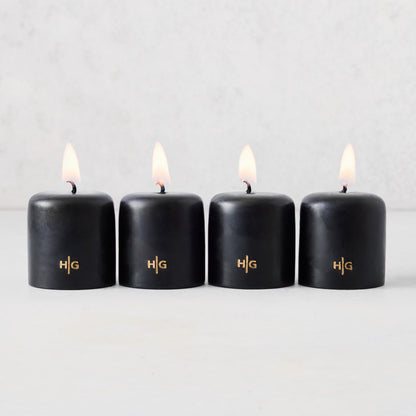 Set of four 2-inch black unscented votive candles with lit wicks and gold embossed HG logo on a light background.