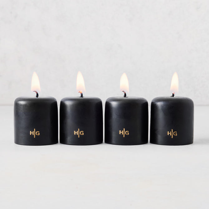 Set of four 2-inch black unscented votive candles with lit wicks and gold embossed HG logo on a light background.