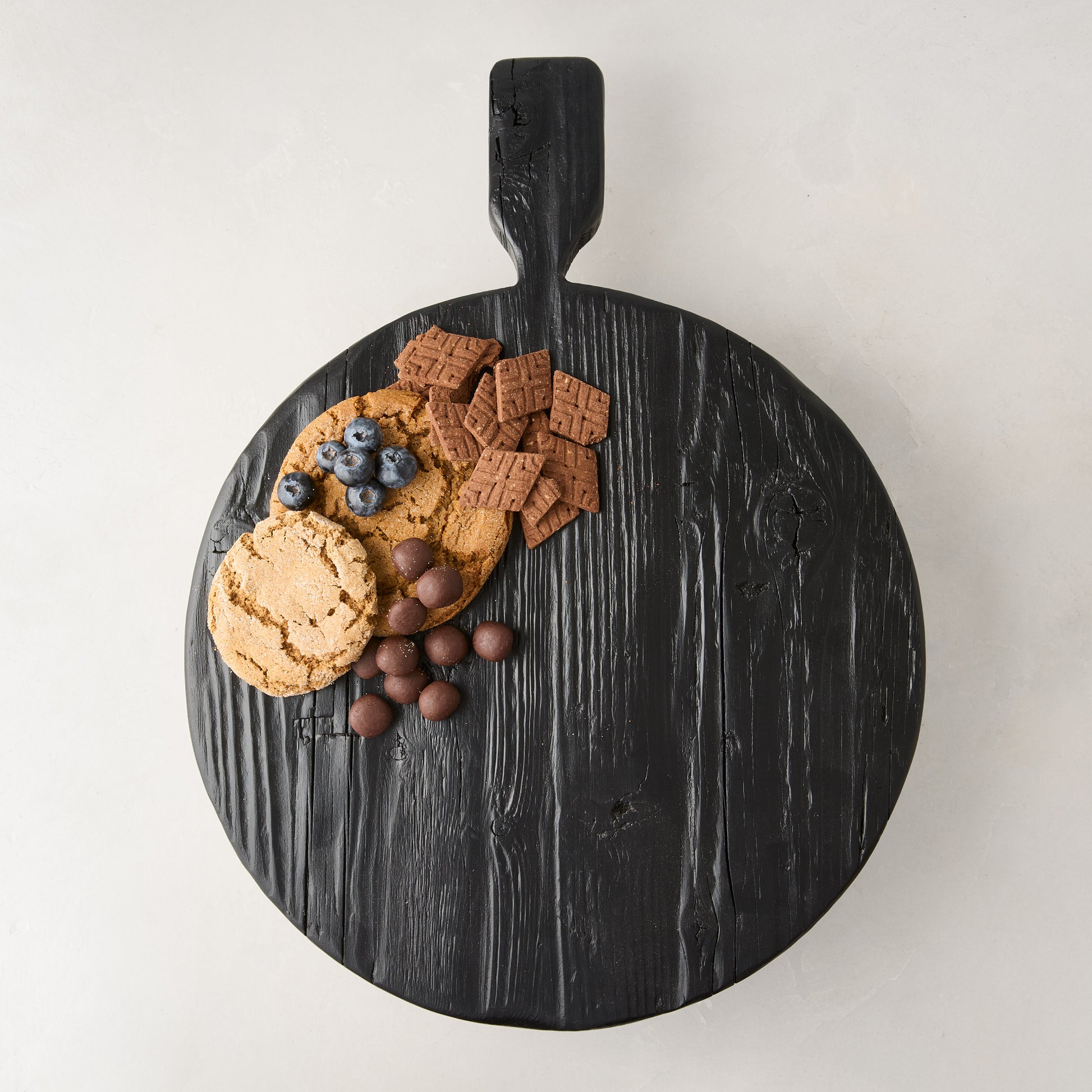 Black wood round footed serve board with handle, displaying cookies, blueberries, and chocolates.