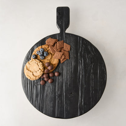 Black wood round footed serve board with handle, displaying cookies, blueberries, and chocolates.