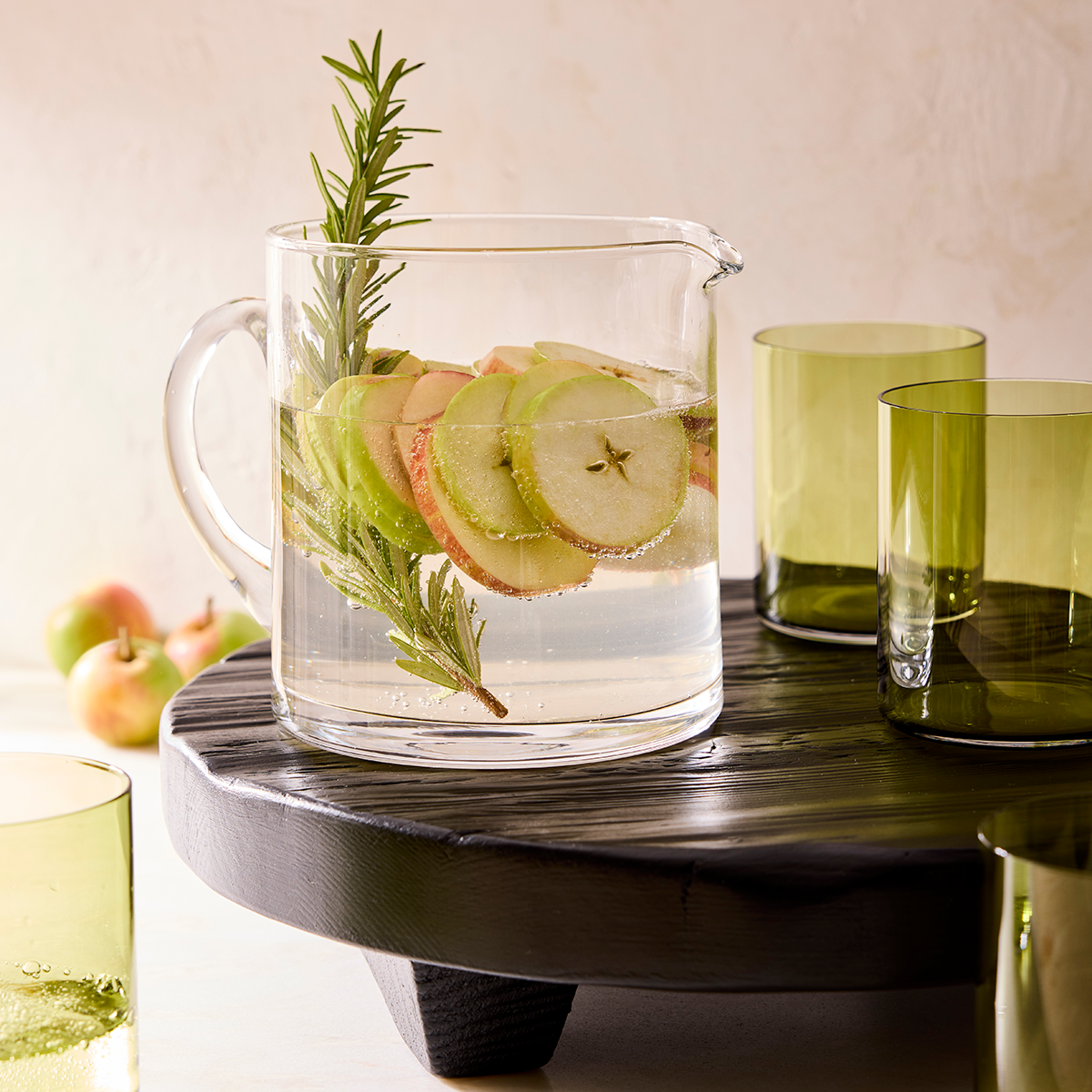 Black wood round footed serve board with handle, holding a pitcher of apple water and green glasses.