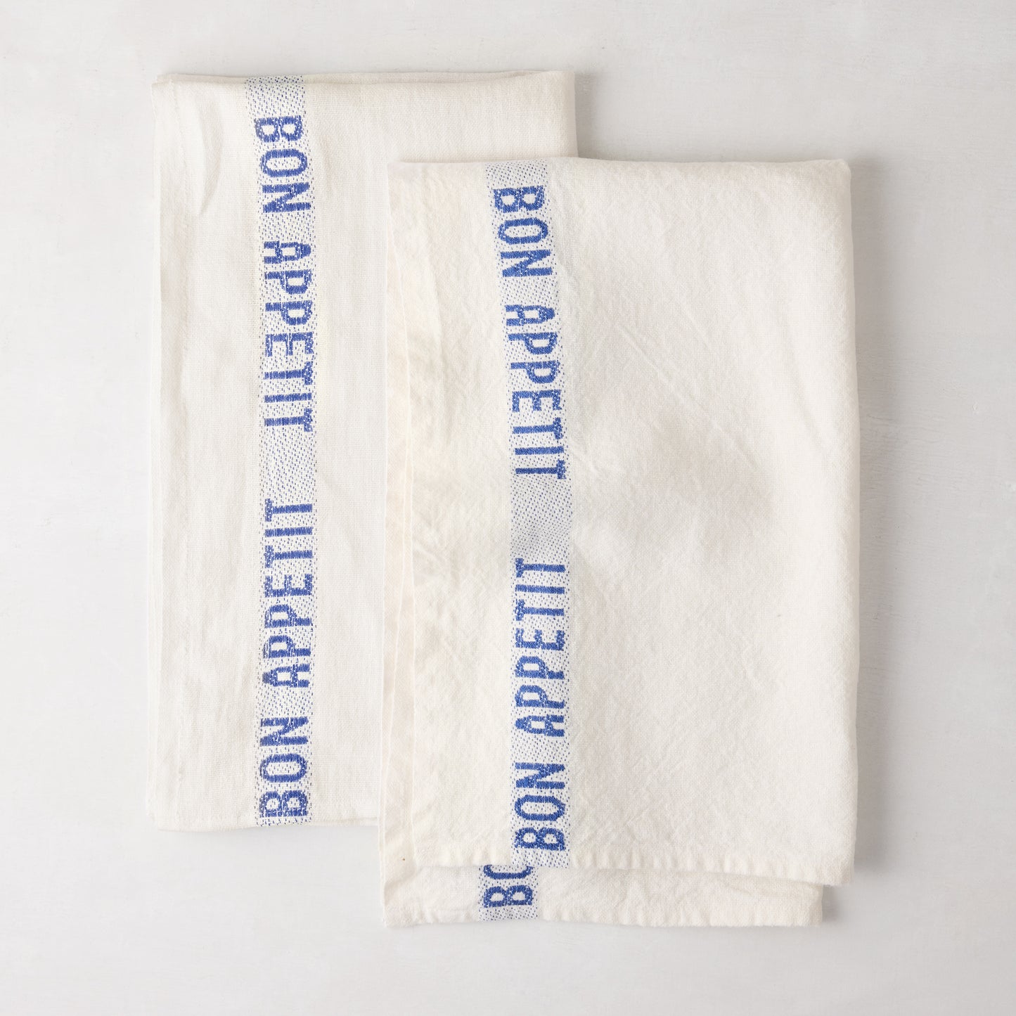 Set of 2 folded white linen hand towels with blue "Bon Appetit" text stripe.