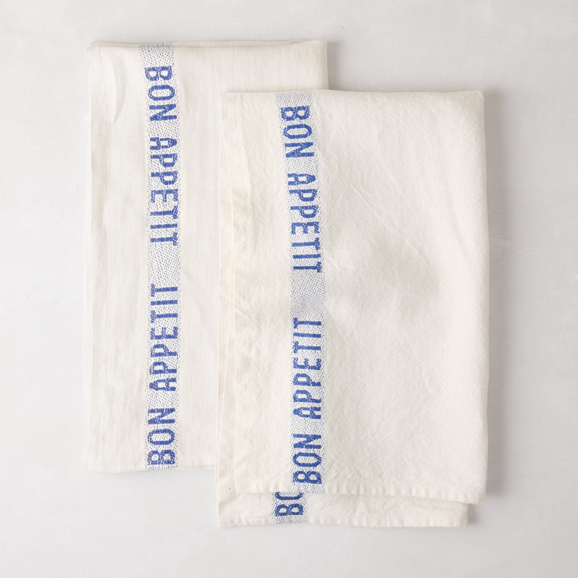 Set of 2 folded white linen hand towels with blue "Bon Appetit" text stripe.