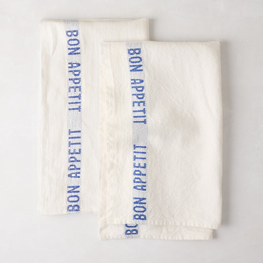 Set of 2 folded white linen hand towels with blue "Bon Appetit" text stripe.