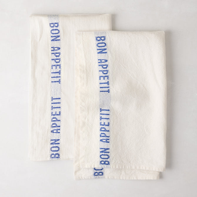 Set of 2 folded white linen hand towels with blue "Bon Appetit" text stripe.