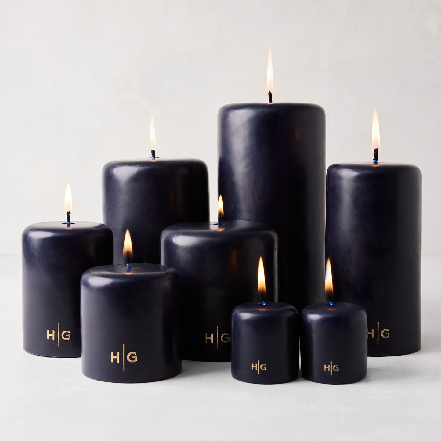 Set of seven lit HG blue unscented paraffin pillar candles in various sizes, with gold HG logo.