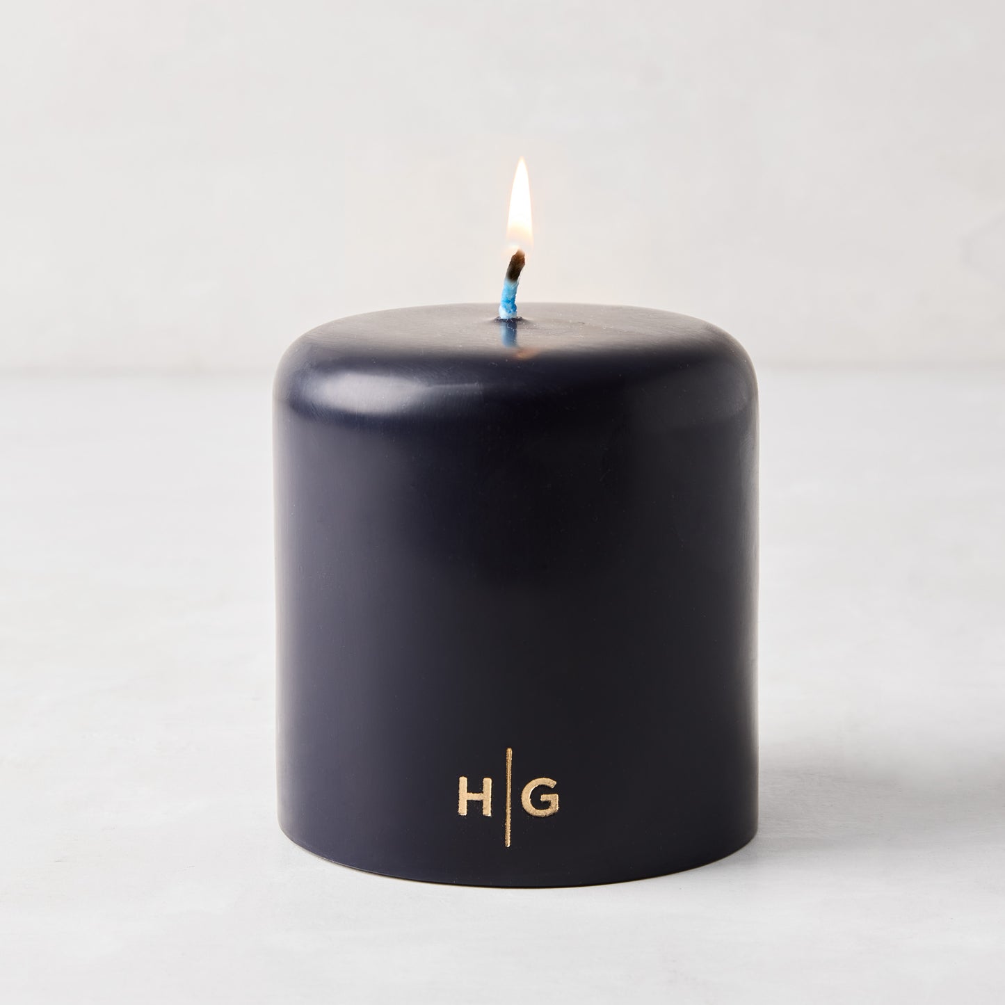 HG Blue unscented 4x4 pillar candle with gold HG logo and lit cotton wick.
