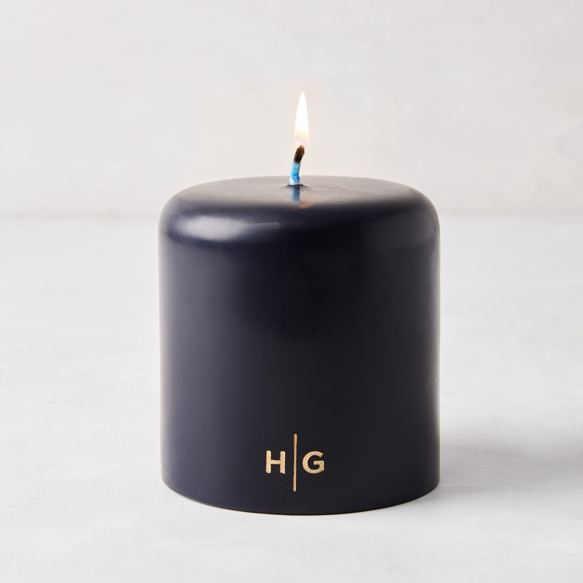 HG Blue unscented 4x4 pillar candle with gold HG logo and lit cotton wick.