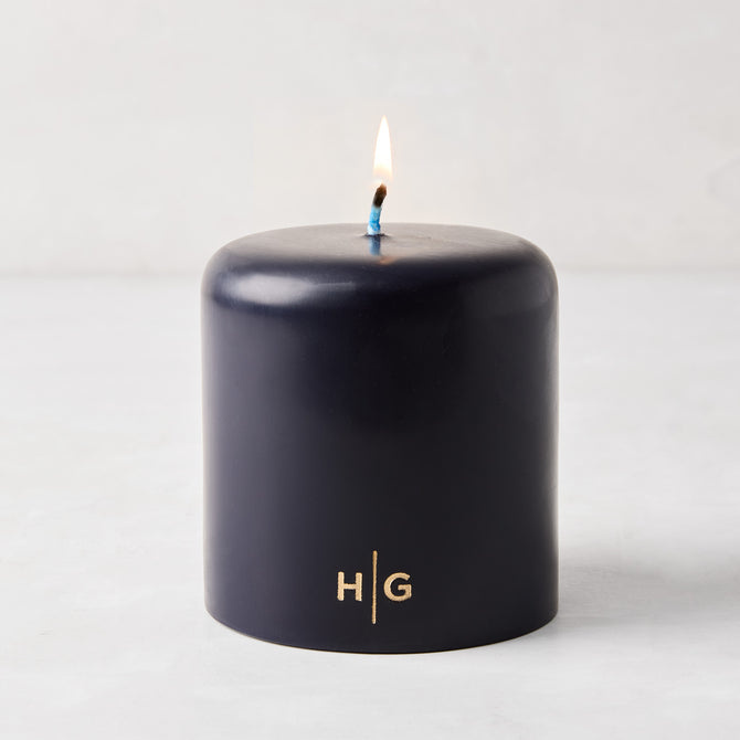 HG Blue unscented 4x4 pillar candle with gold HG logo and lit cotton wick.