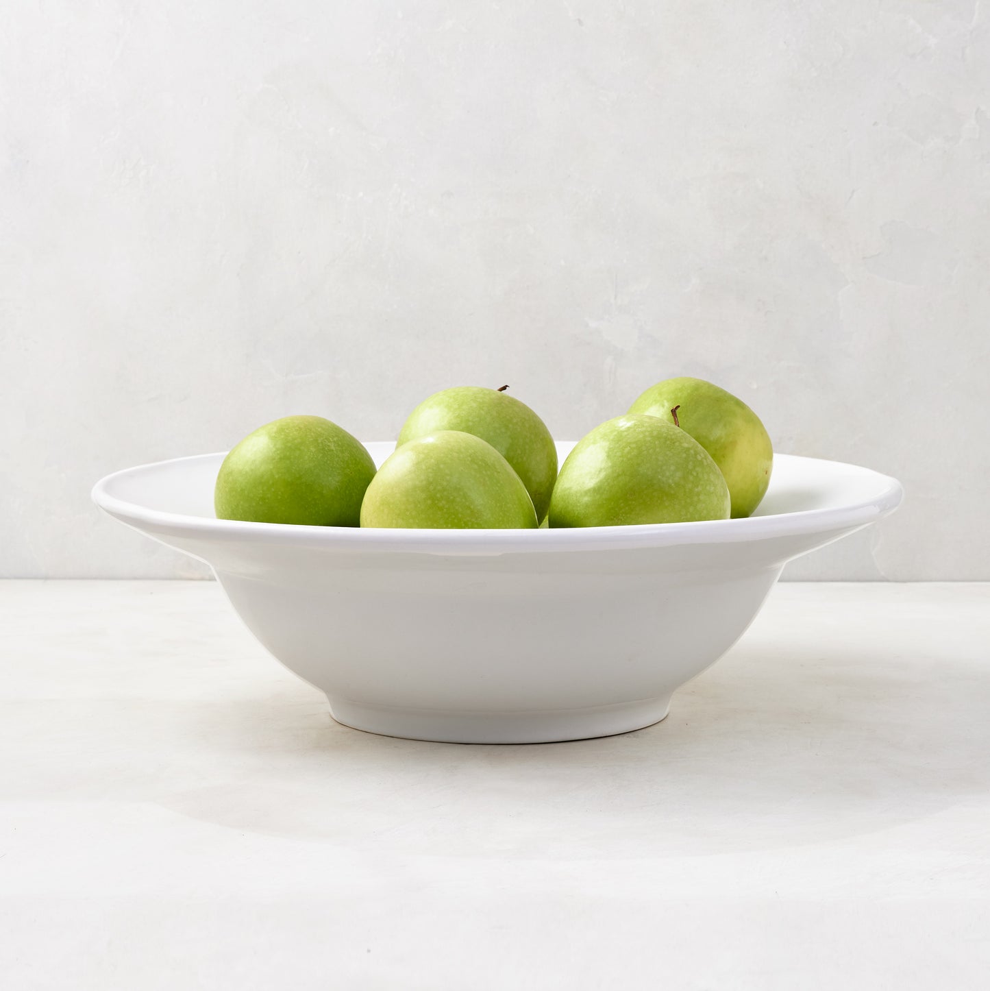White ceramic Bolinas round serve bowl with raised trim, filled with green apples.