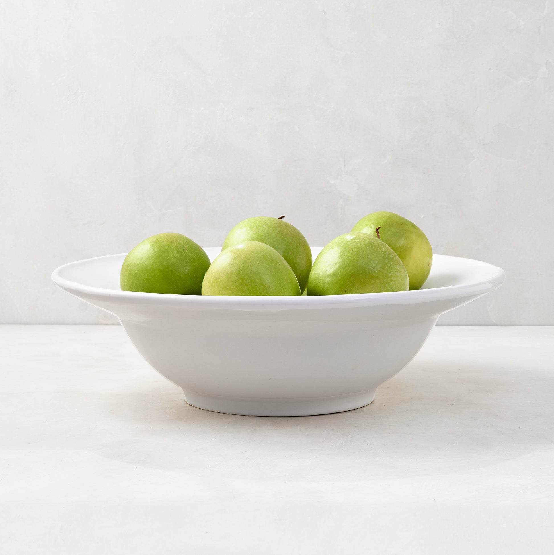 White ceramic Bolinas round serve bowl with raised trim, filled with green apples.