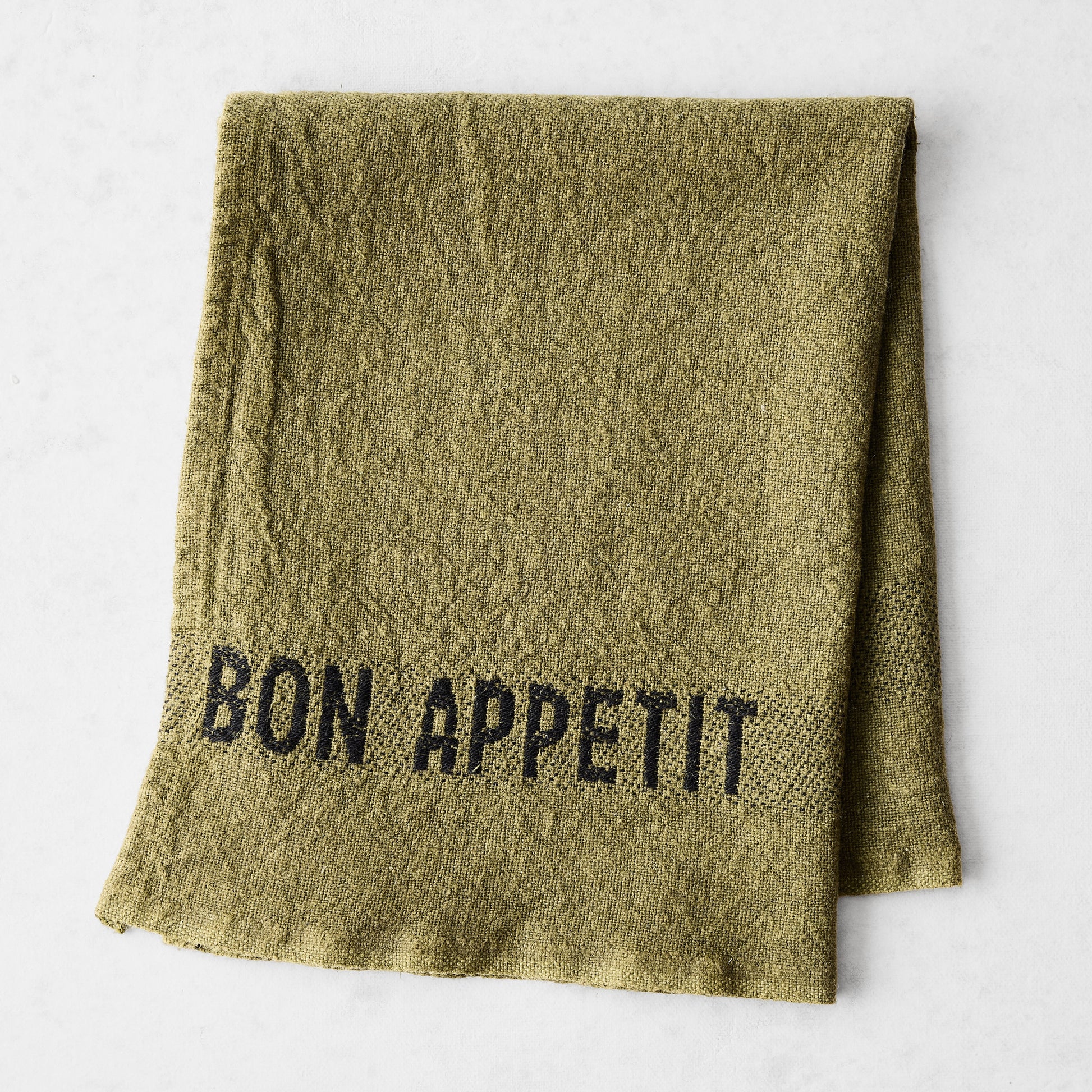 Folded avocad green linen napkin with "BON APPETIT" in black lettering.