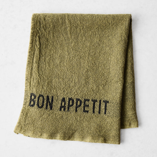 Folded avocad green linen napkin with "BON APPETIT" in black lettering.