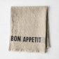 Camel khaki linen napkin with "BON APPETIT" in black lettering, folded on a white surface.