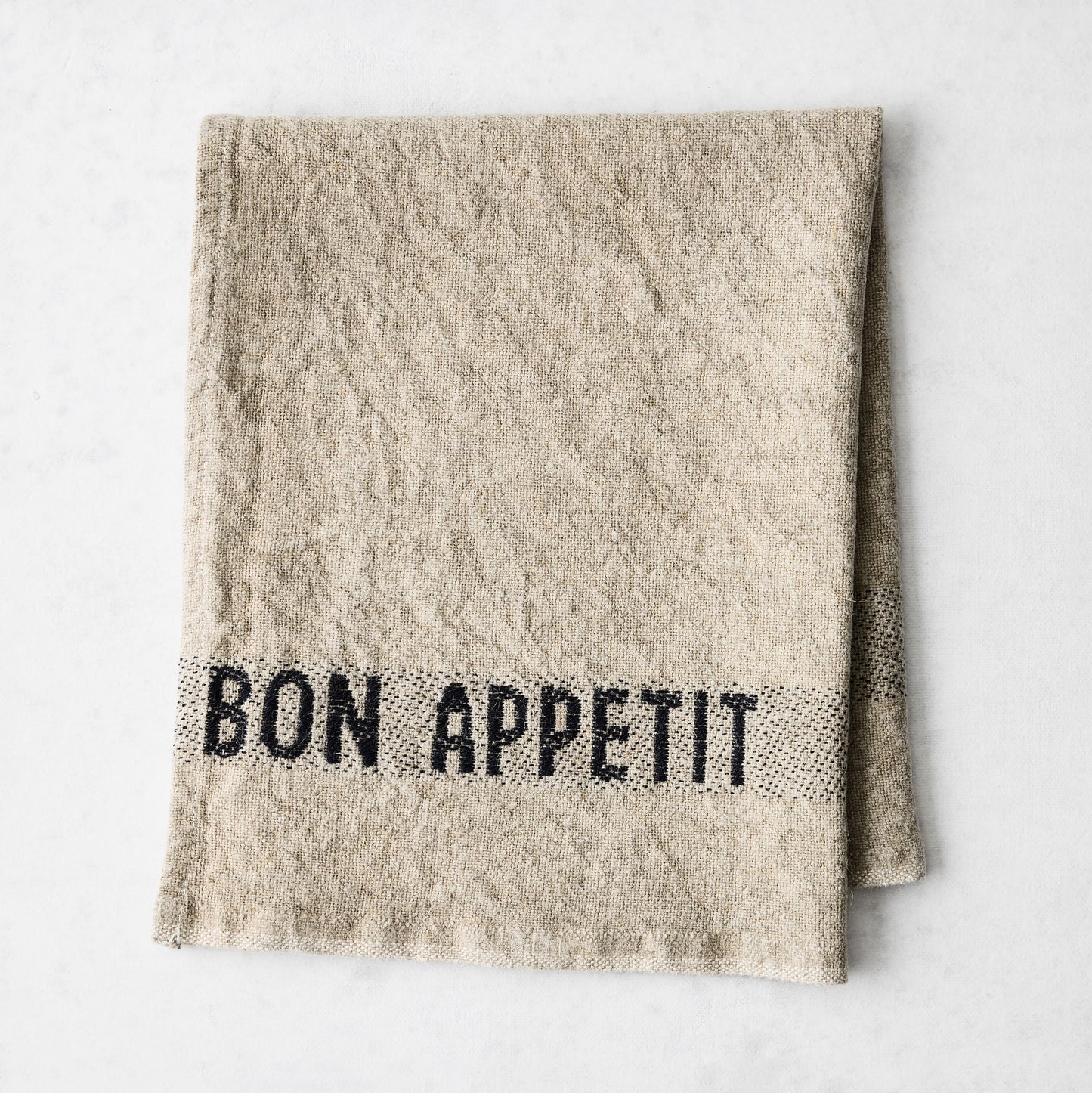 Camel khaki linen napkin with "BON APPETIT" in black lettering, folded on a white surface.