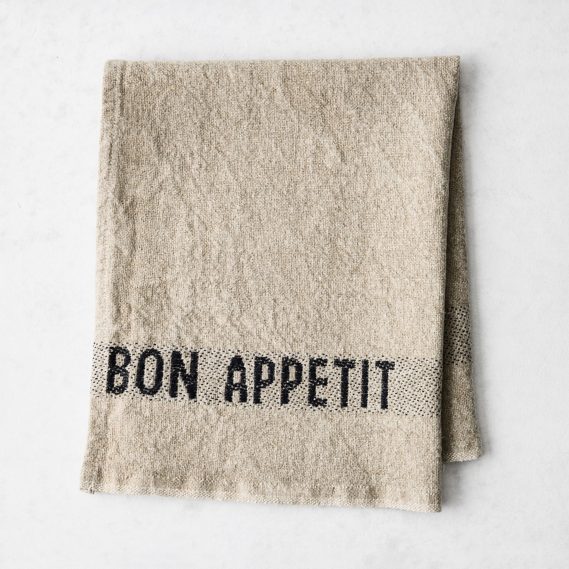Camel khaki linen napkin with "BON APPETIT" in black lettering, folded on a white surface.