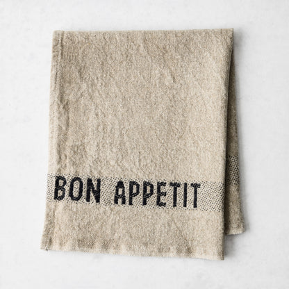 Camel khaki linen napkin with "BON APPETIT" in black lettering, folded on a white surface.
