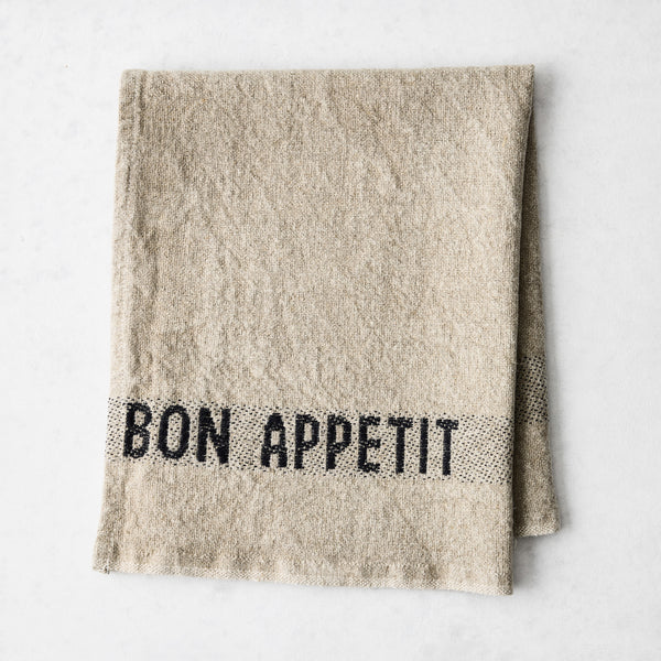 Camel khaki linen napkin with "BON APPETIT" in black lettering, folded on a white surface.