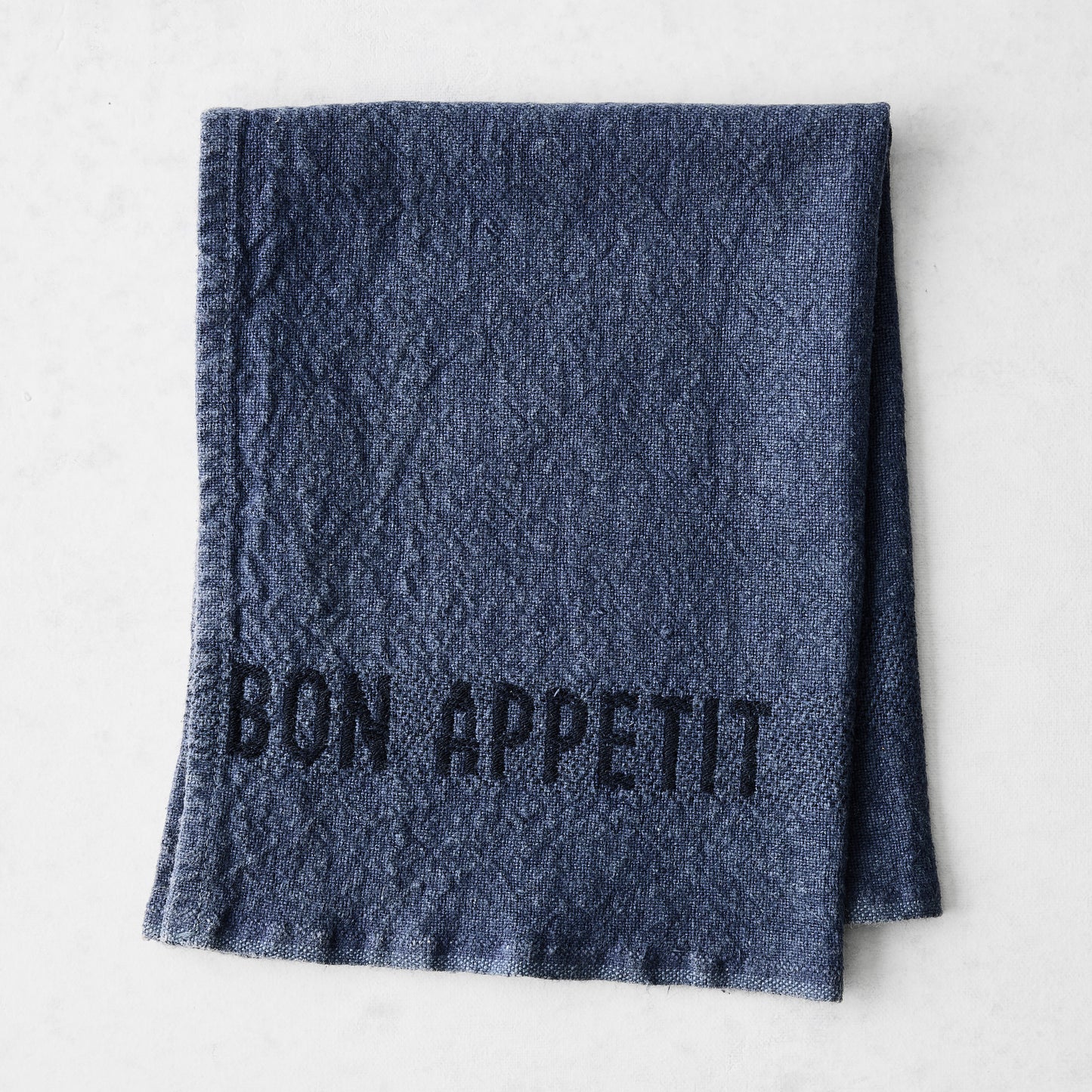 Indigo blue linen napkin with "BON APPETIT" in black lettering, folded on a white surface.