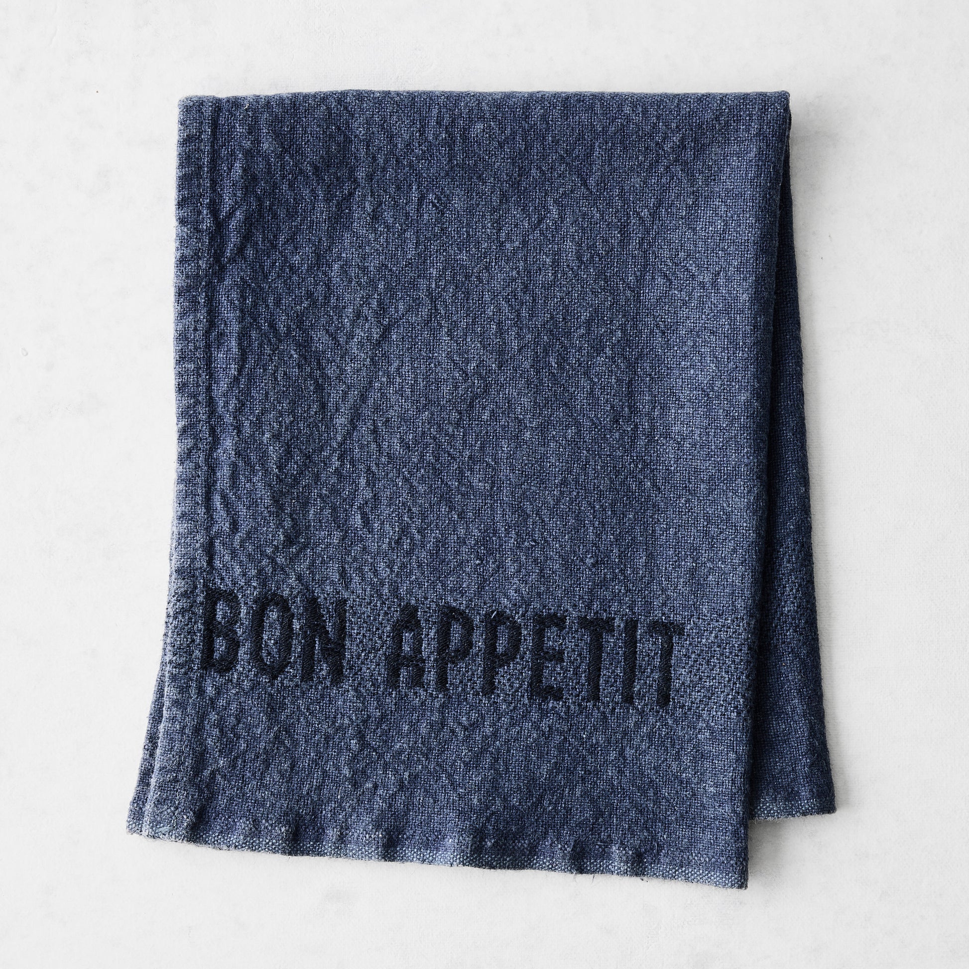 Indigo blue linen napkin with "BON APPETIT" in black lettering, folded on a white surface.