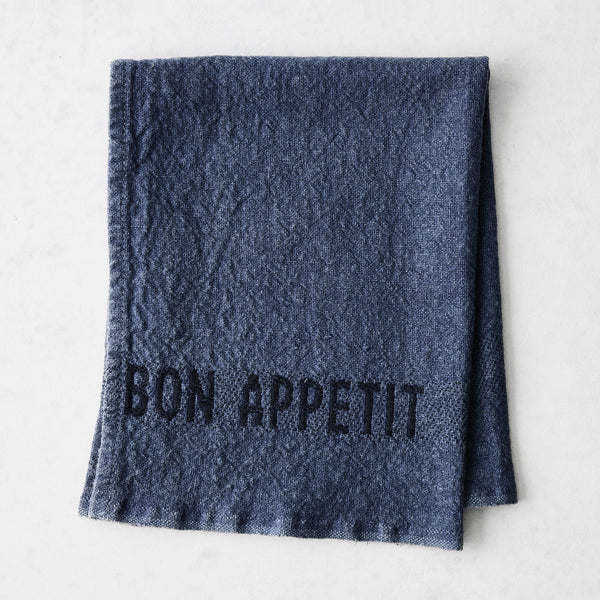 Indigo blue linen napkin with "BON APPETIT" in black lettering, folded on a white surface.